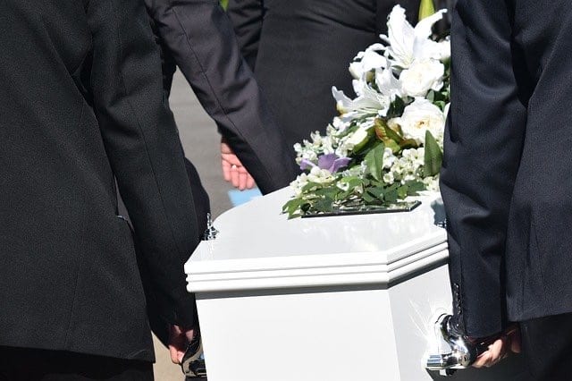 A white casket is carried with purple flowers